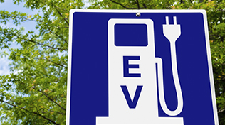 Close photo of a Blue Sign indicating an Electric Vehicle Recharging Point. Green Trees are Visible in Background. Ecological Mode of Transport.